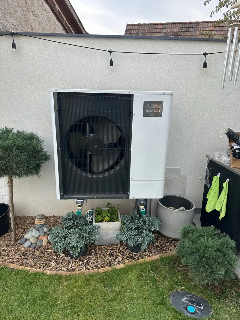 Mitsubishi Electric levegő-víz hőszivattyú kültéri egysége egy rendezett kert sarkában, örökzöldek között – Vác